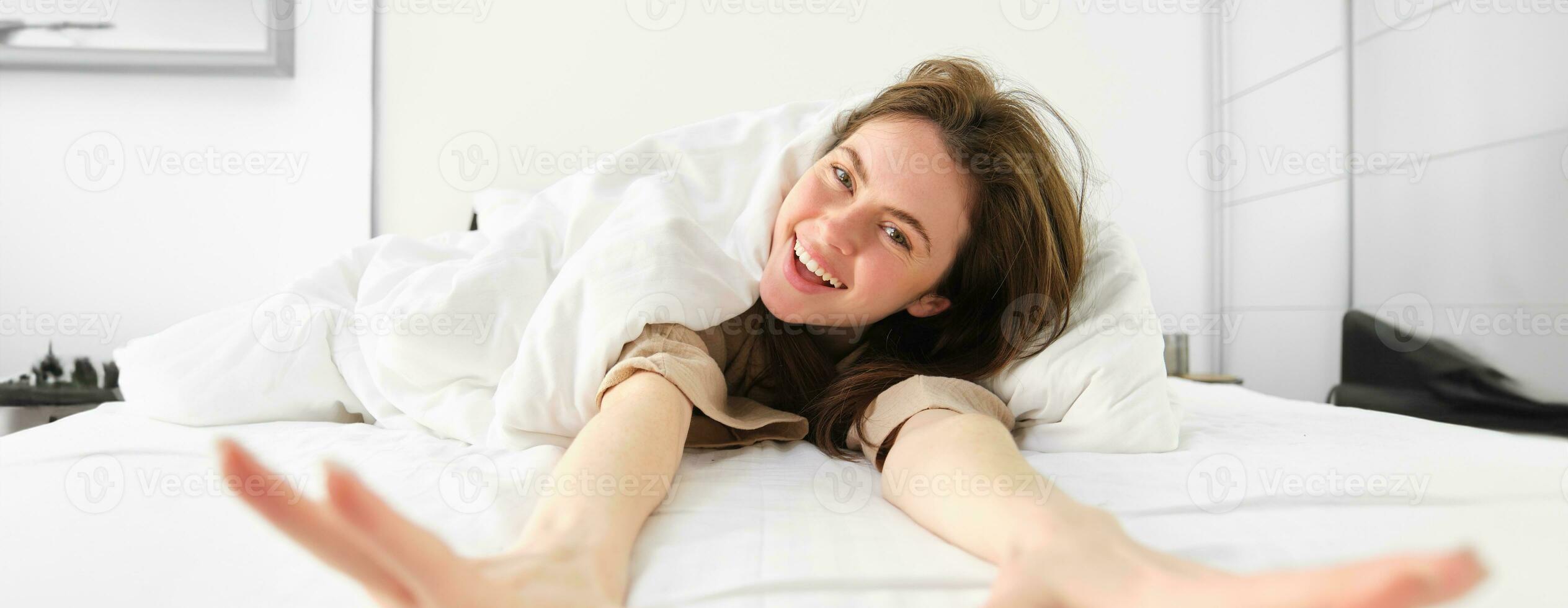 retrato do feliz jovem mulher acordando acima enérgico dentro manhã, alongamento braços para Câmera e rindo dentro dela cama debaixo cobertores foto