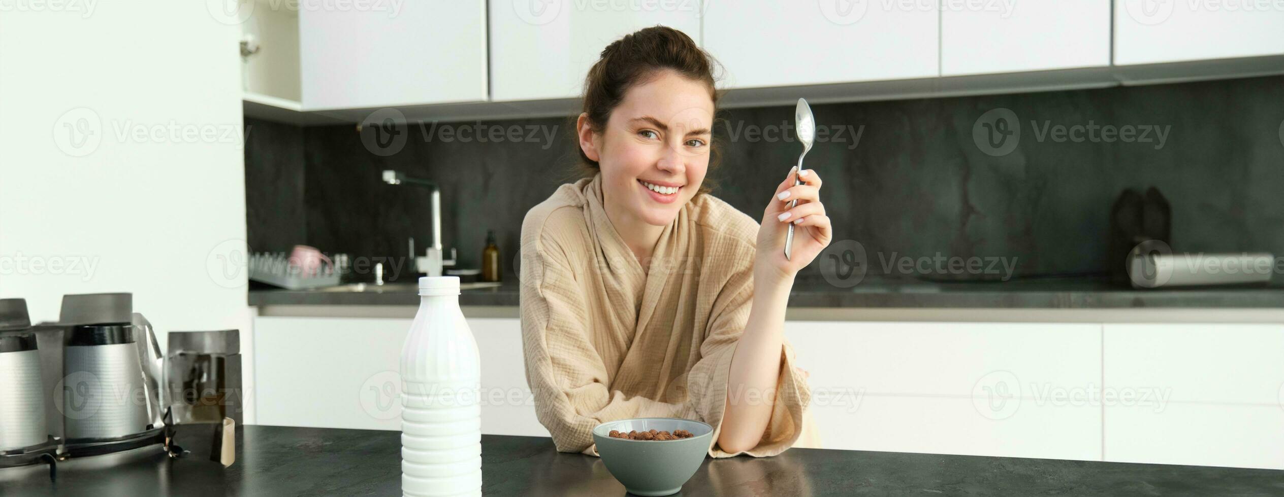 retrato do lindo jovem e saudável mulher dentro roupão de banho come dela café da manhã dentro cozinha, tem cereais com leite e sorridente foto