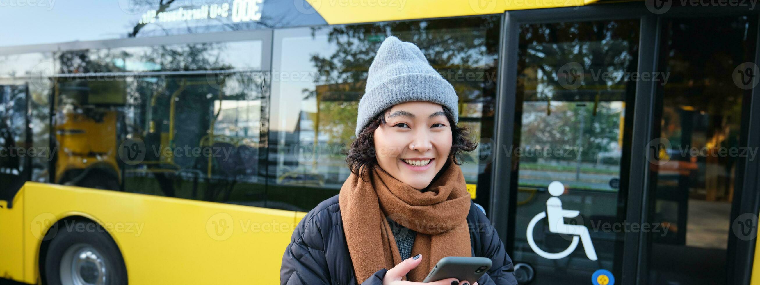 imagem do menina aluna esperando para público transporte, Verificações cronograma em Smartphone aplicativo, carrinhos perto cidade ônibus foto