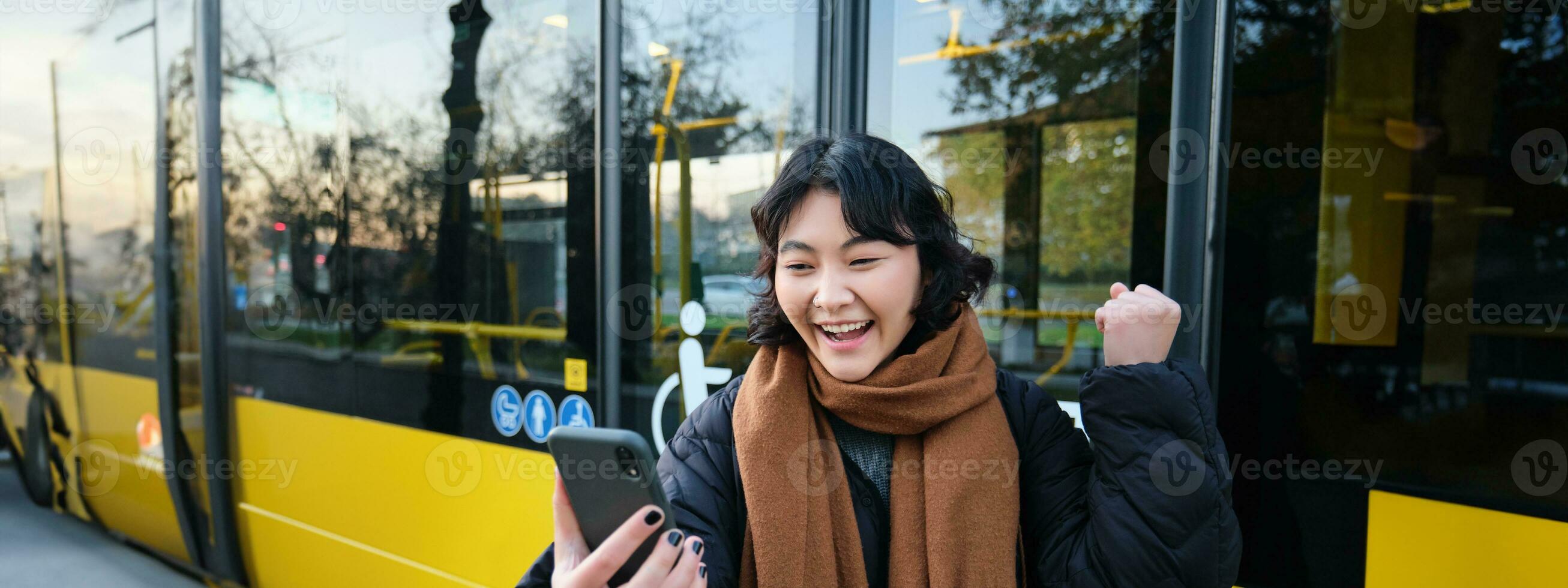 retrato do alegre ásia menina fala em Móvel telefone, vídeo bate-papos, parece espantado às Smartphone Câmera, carrinhos em ônibus Pare foto