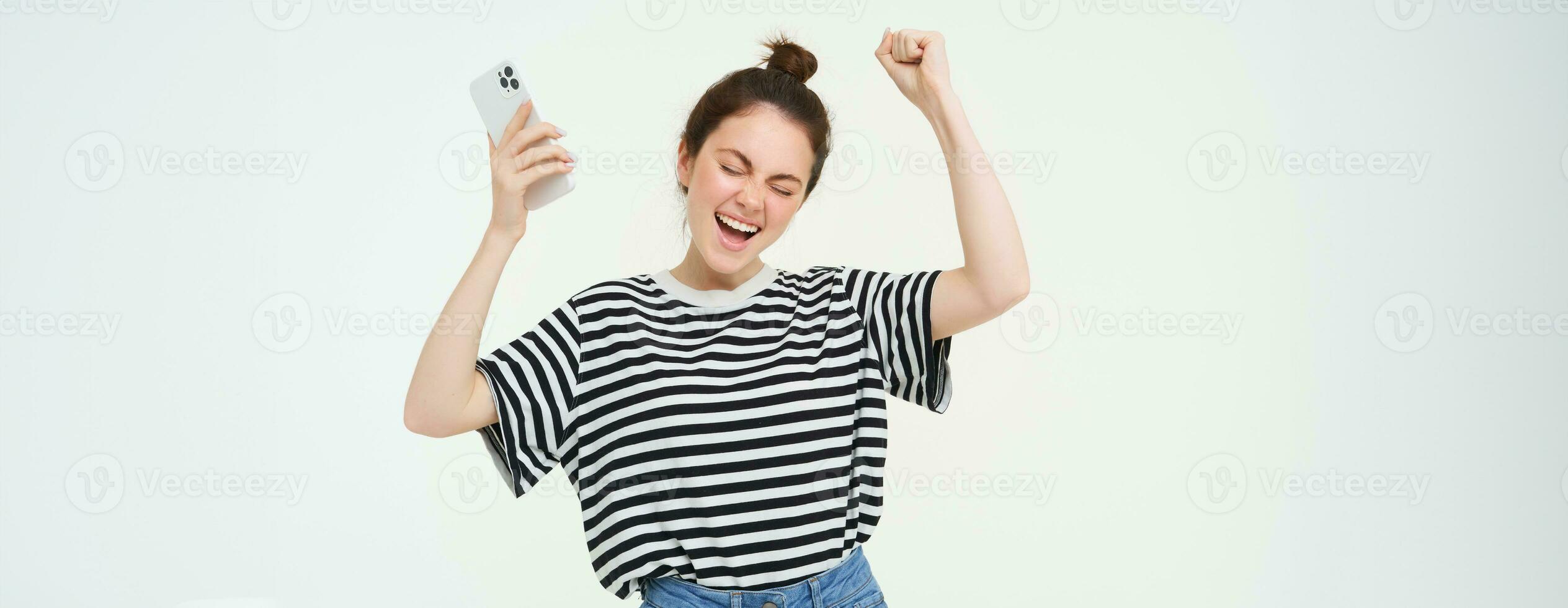 menina dançando com Móvel telefone contra branco fundo. mulher com Smartphone cantando, levantando mãos acima e a comemorar foto