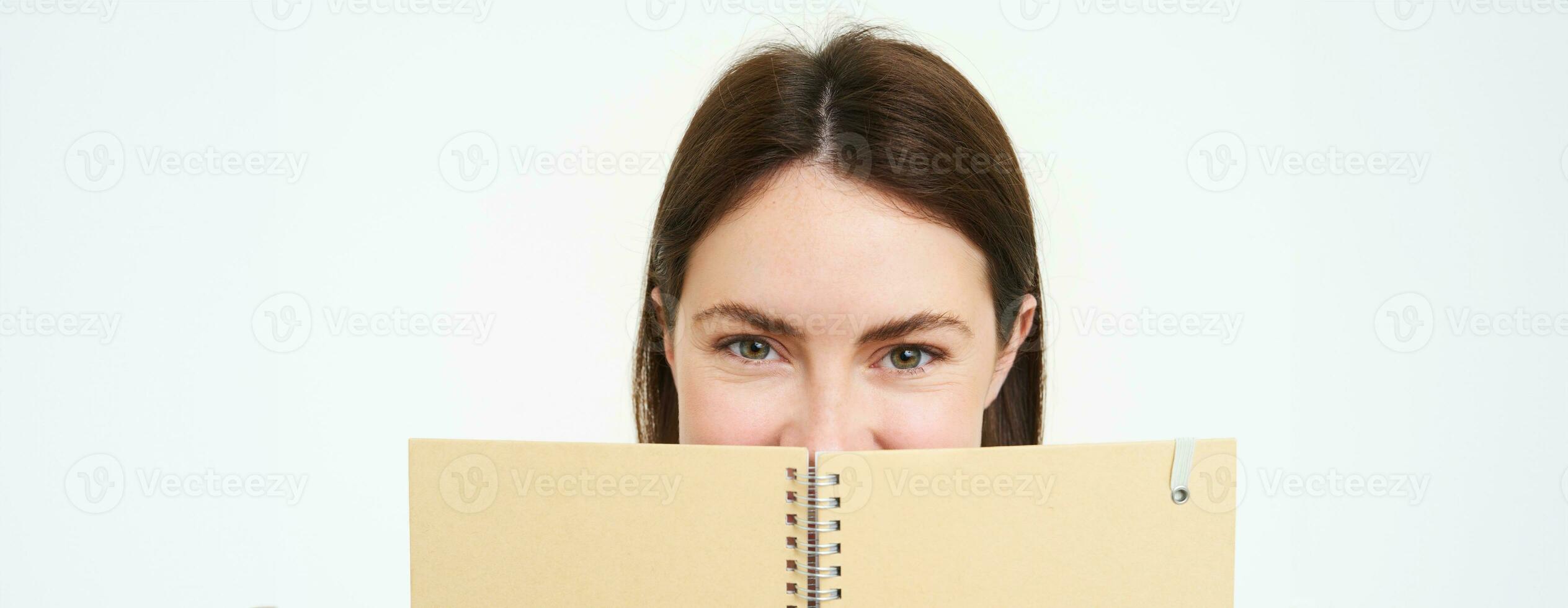 retrato do fofa jovem mulher esconde dela face atrás planejador, detém caderno contra dela face e sorrisos, isolado sobre branco fundo foto