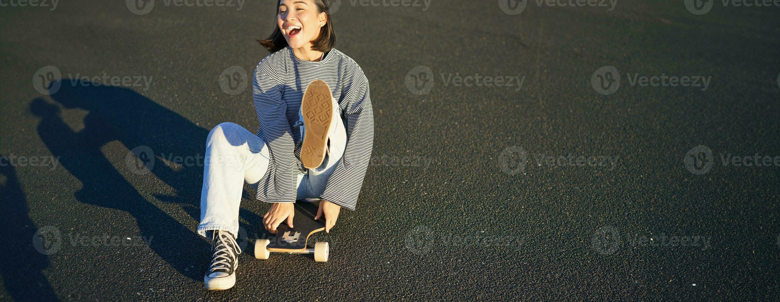 retrato do lindo ásia menina patinação, sentado em dela skate e sorridente. fofa adolescente com Longboard, patinação em estrada foto