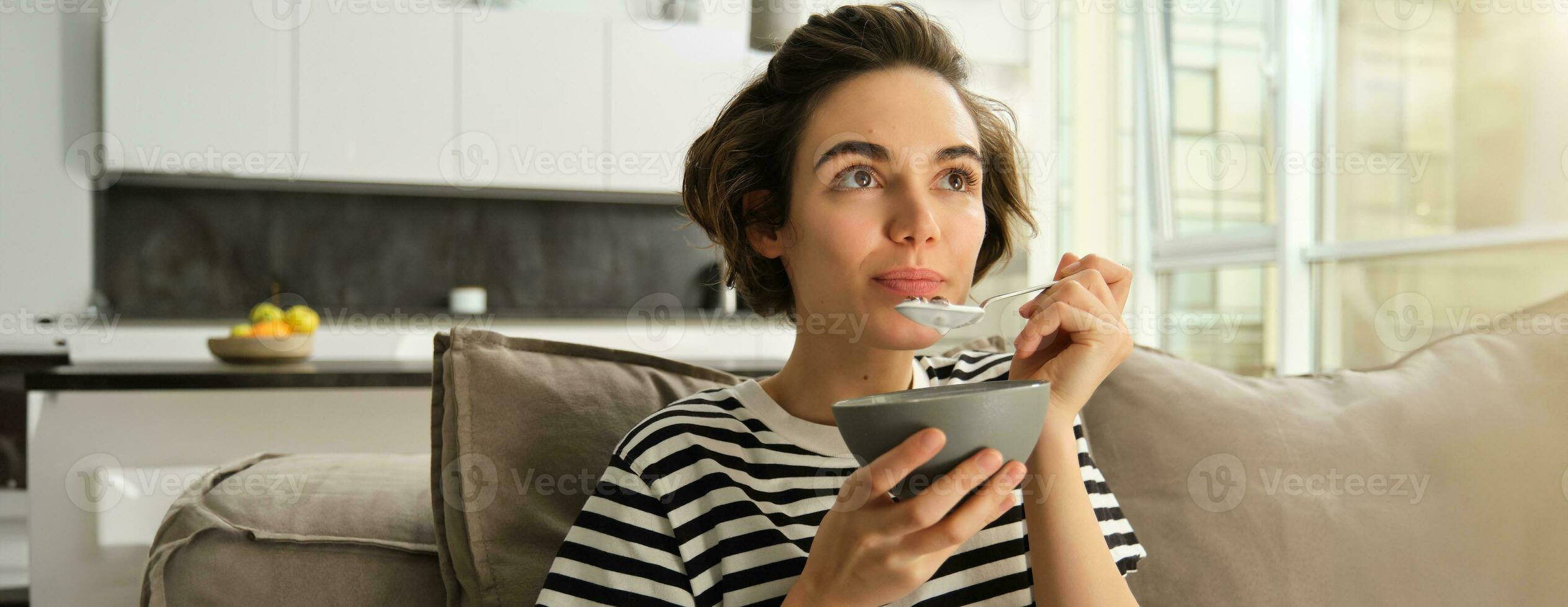 retrato do sorridente fêmea modelo, comendo cereais com leite, tendo café da manhã dentro frente do televisão dentro vivo sala, assistindo televisão foto