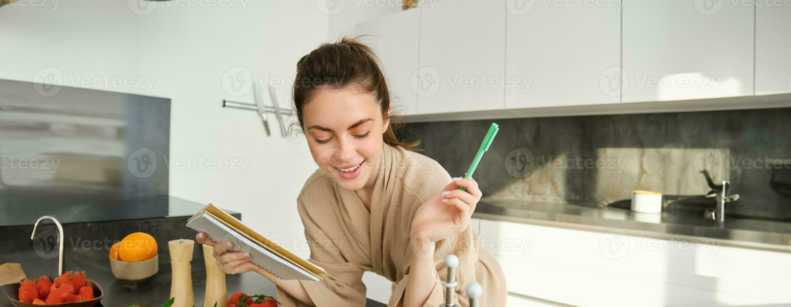 retrato do mulher verificação mercearia lista, olhando às vegetais, segurando caderno, lendo receita enquanto cozinhando refeição dentro a cozinha, cortar tomates e abobrinha foto