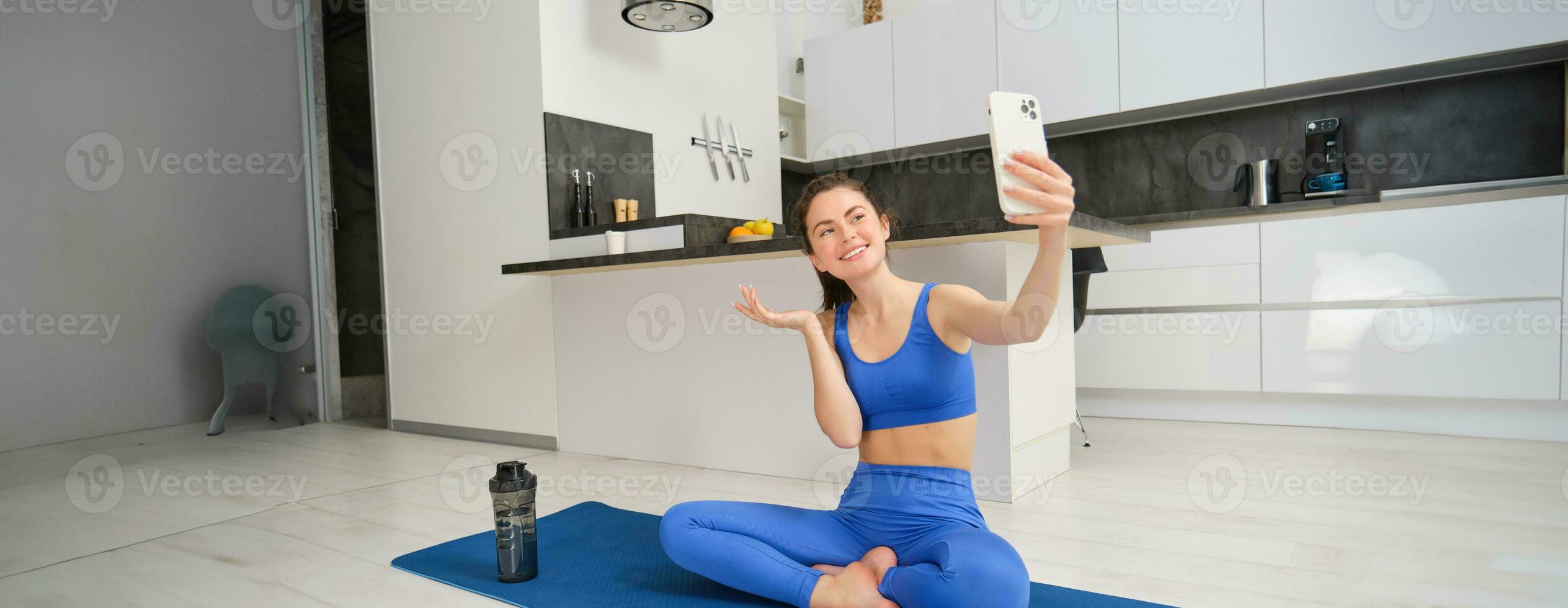 ativo jovem mulher faz Esportes, registros dela exercite-se Treinamento a partir de casa em Smartphone Câmera, posando para selfie dentro dela casa, senta em borracha ioga esteira dentro azul perneiras e sportsbra foto