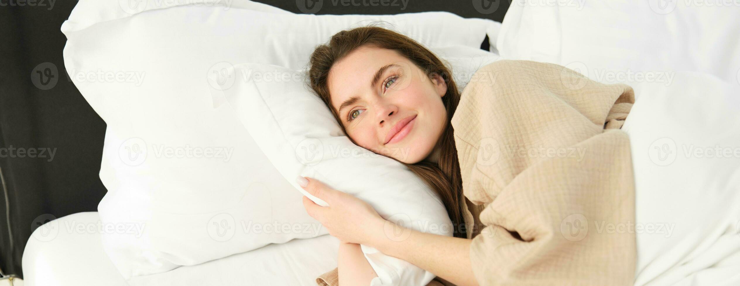 linda mulher em repouso dentro cama vestindo pijamas, abraçando dela travesseiro e sorridente foto