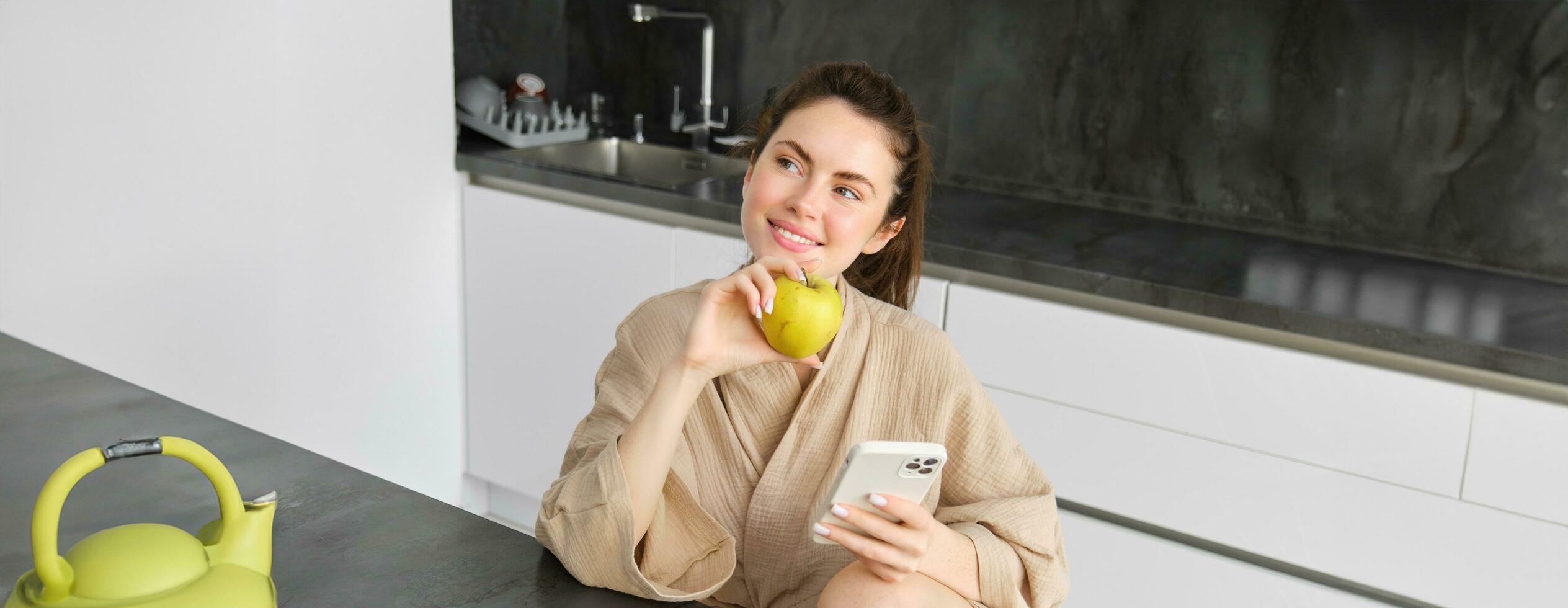 imagem do atraente jovem mulher dentro roupão de banho, senta dentro cozinha, come verde maçã e parece às Móvel telefone, usa Smartphone aplicativo, ordens Comida em inscrição foto