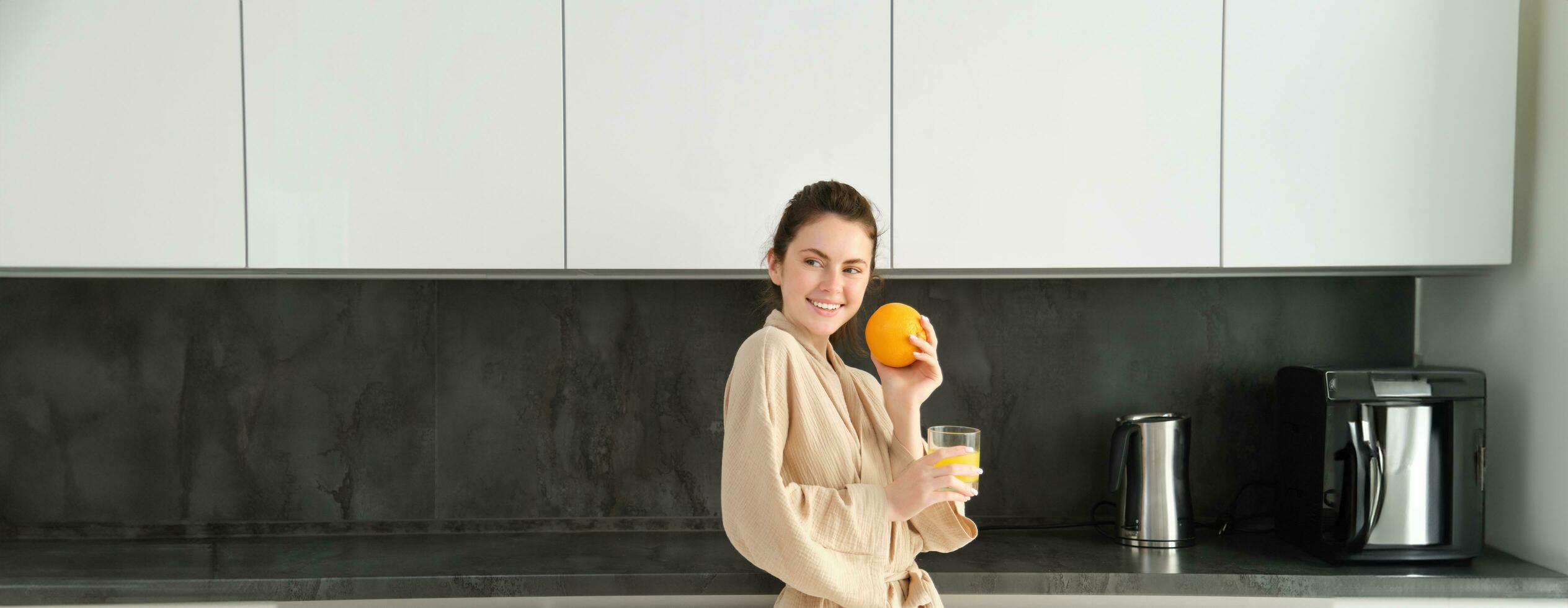 retrato do jovem e saudável mulher começa dela dia com frutas, comendo fresco laranja, bebendo suco dentro cozinha, vestindo roupão de banho foto