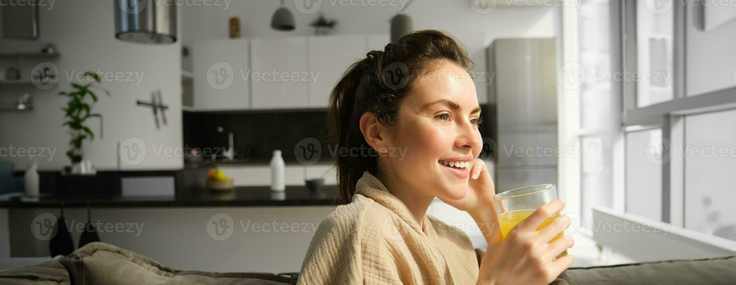 fechar acima do lindo jovem mulher em sofá, bebidas laranja suco e sorrisos, parece lado de fora janela, goza manhã às casa foto