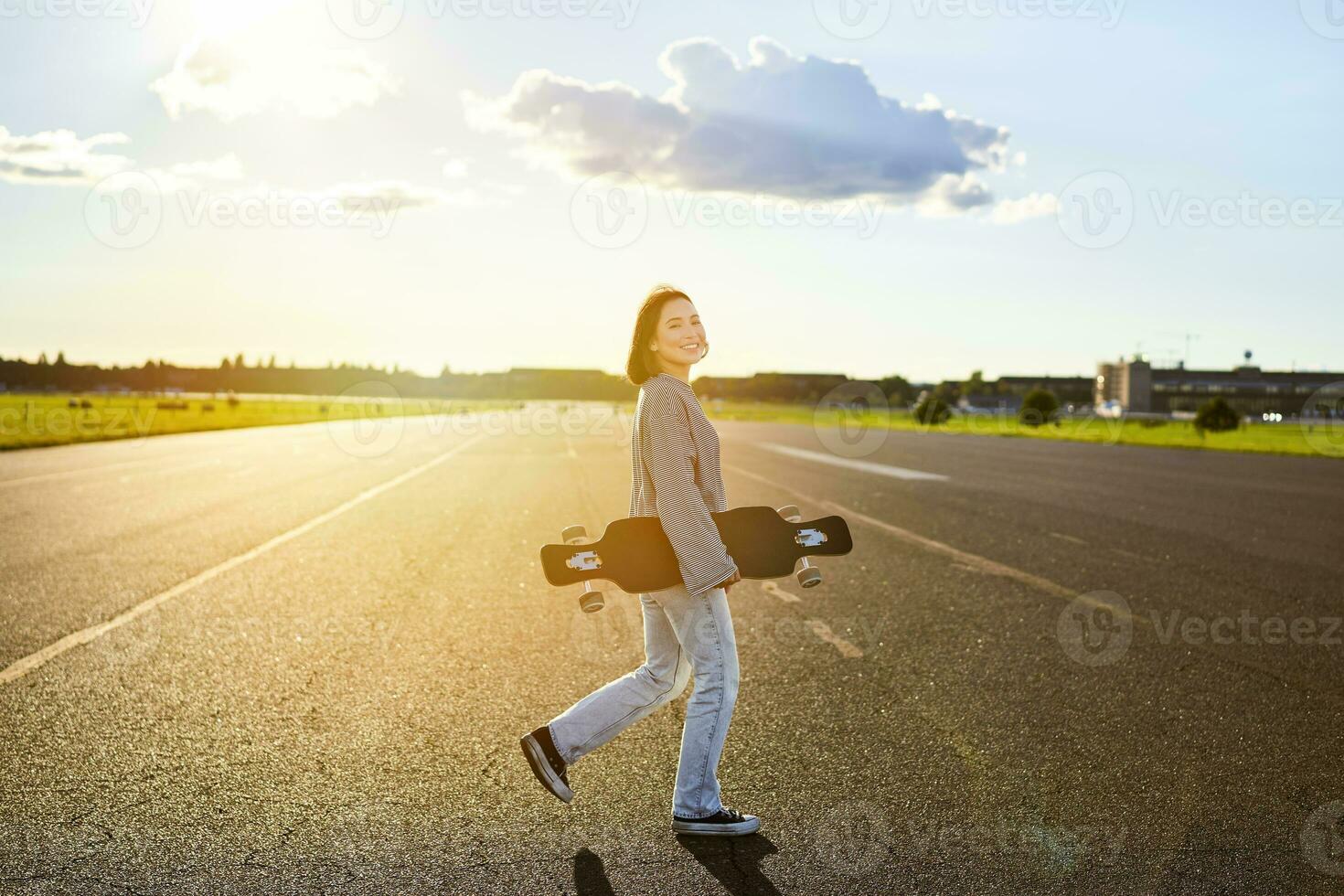 ásia menina com skate em pé em estrada durante pôr do sol. skatista posando com dela grandes quadro, cruzador área coberta durante Treinamento foto