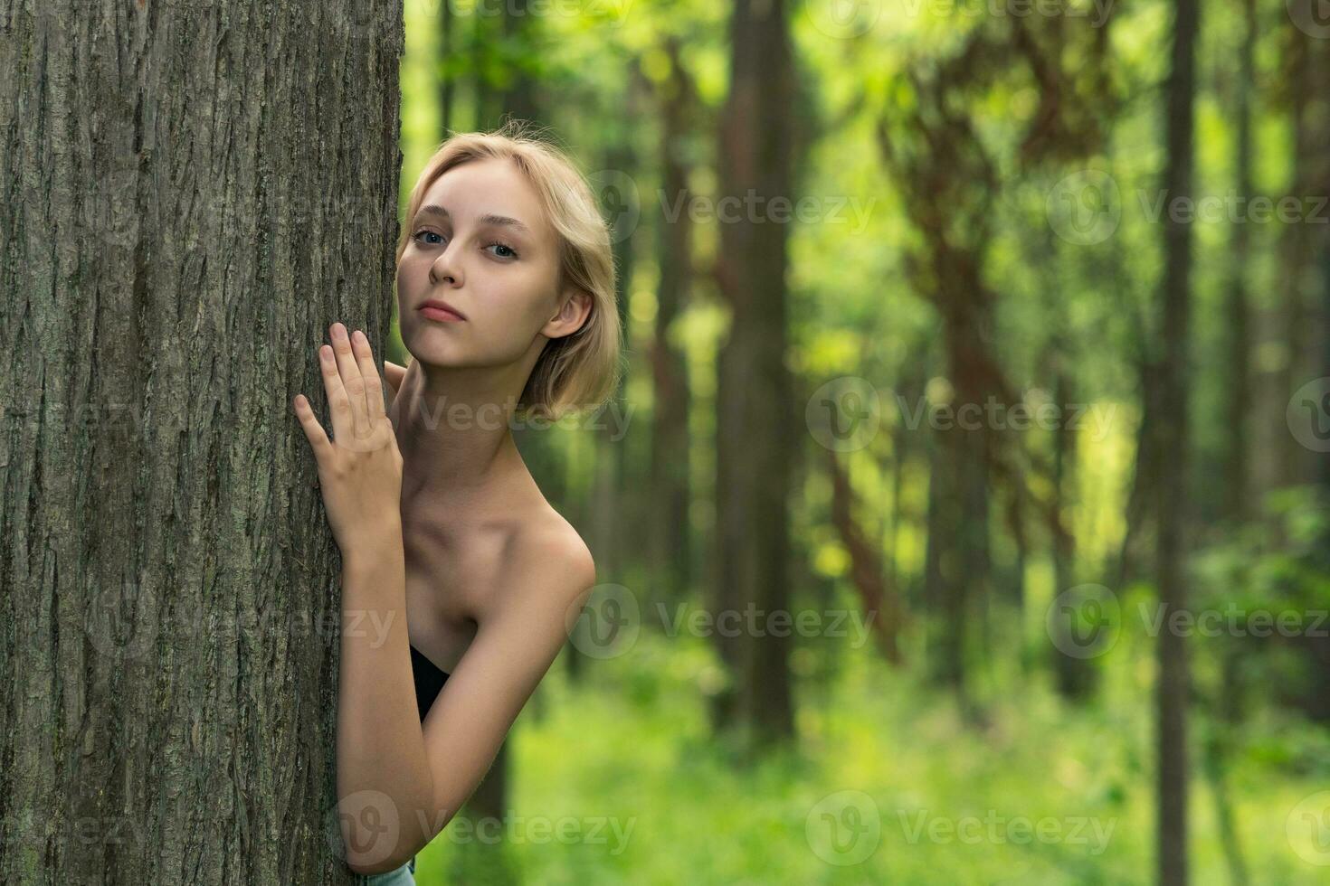 lindo menina espreitar Fora a partir de atrás uma árvore dentro a floresta foto