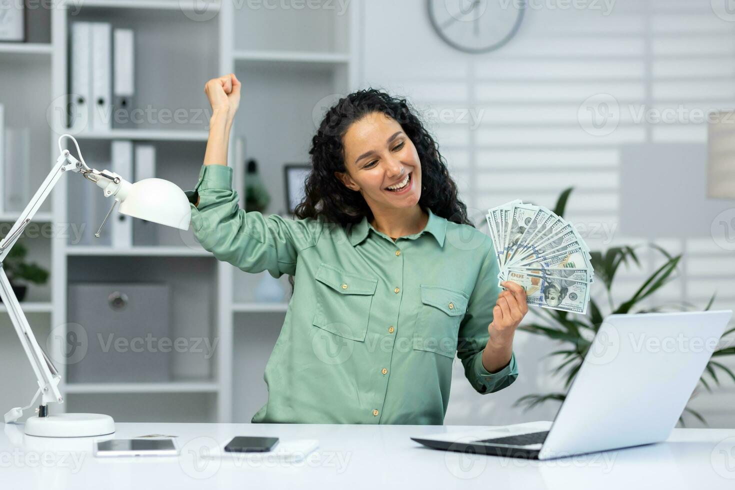 uma jovem hispânico mulher é sentado às a escrivaninha dentro a escritório e segurando dinheiro dentro dela mãos. feliz e sorridente. comemora, danças. foto