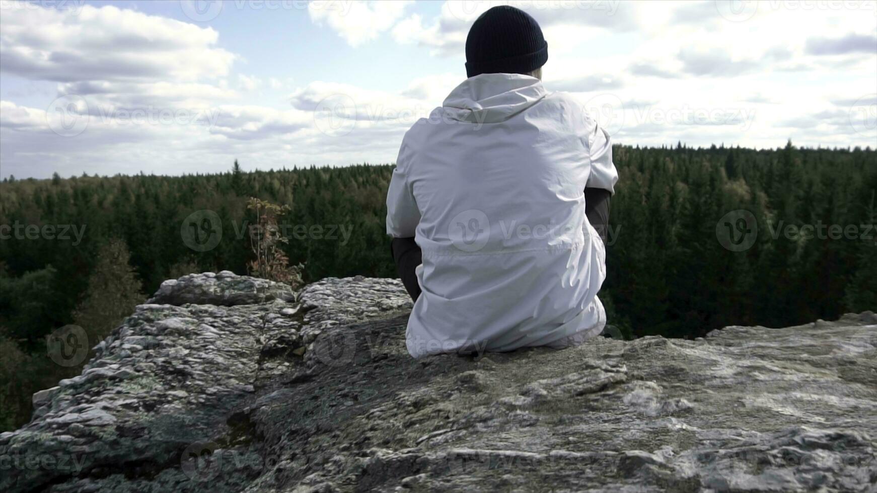 sozinho homem sentado em uma Alto montanha topo olhando às uma nublado céu e árvores picos. traseiro Visão do uma homem dentro a chapéu e branco Jaqueta sentado em uma pedra, olhando às lindo floresta. foto