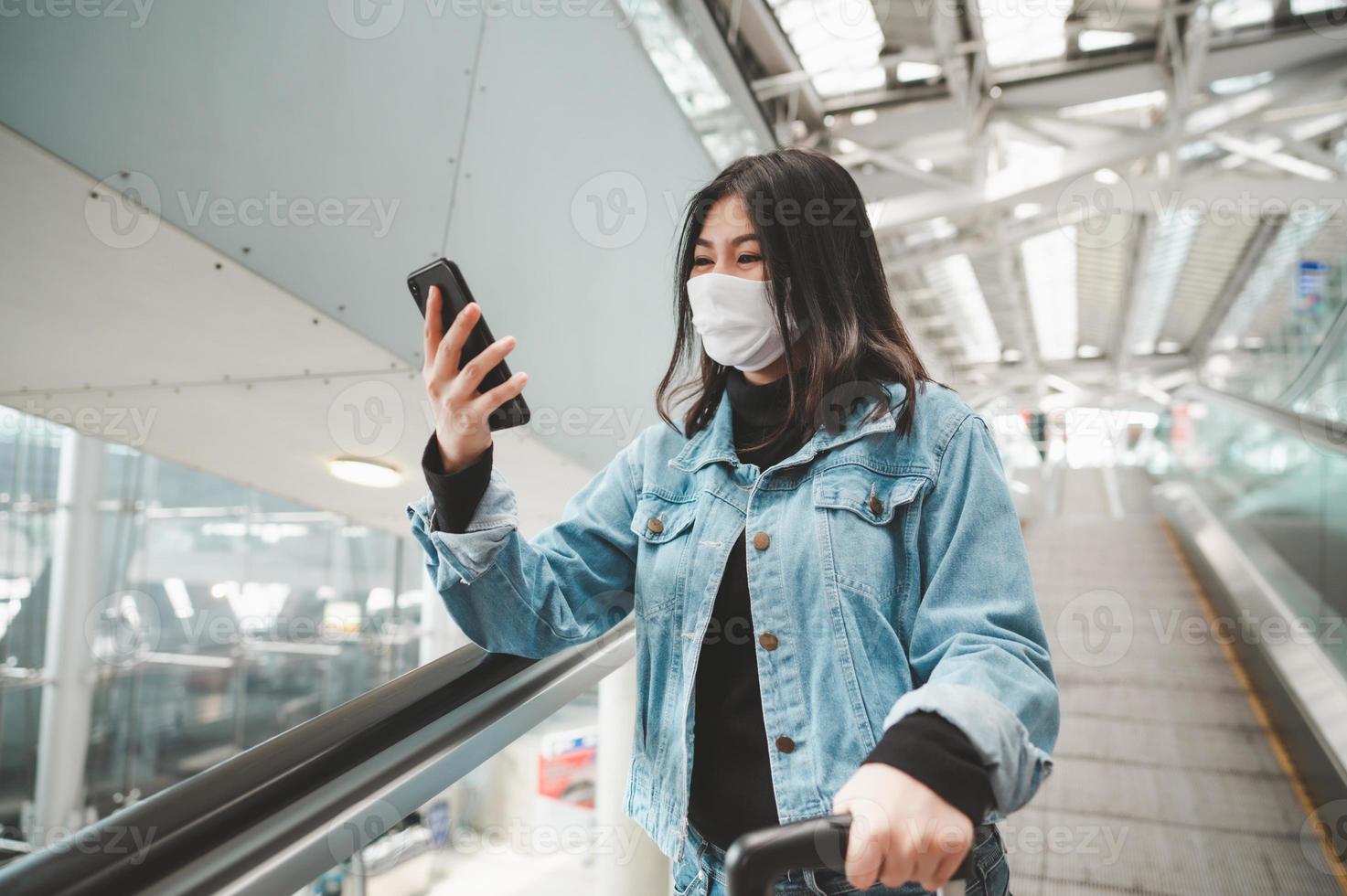 viajante asiática usando máscara usando smartphone em pé na escada rolante foto