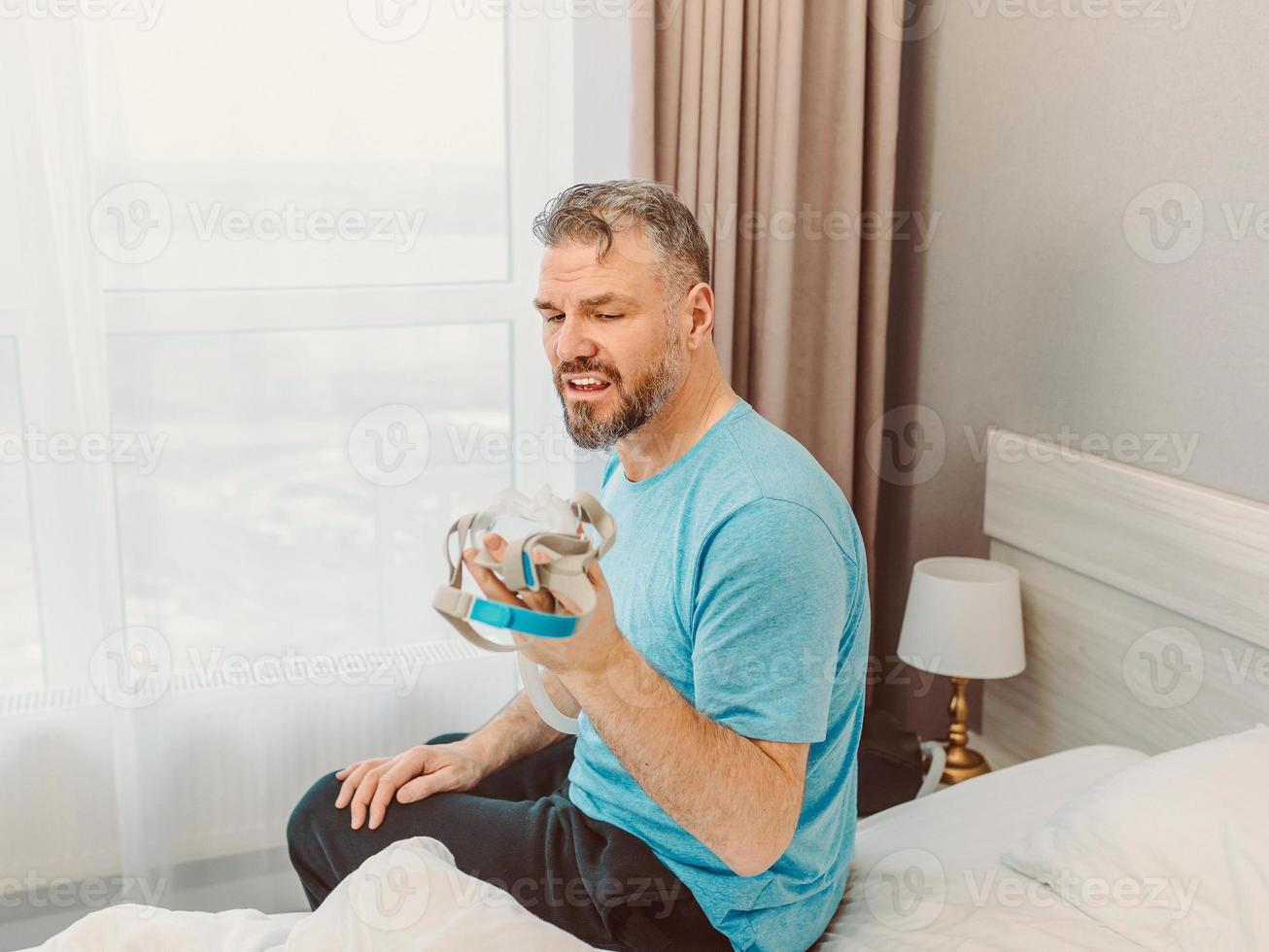homem maduro e feliz com problemas respiratórios crônicos, considera o uso de sentar na cama no quarto com a máquina de cpap. saúde, cpap, conceito de ronco foto