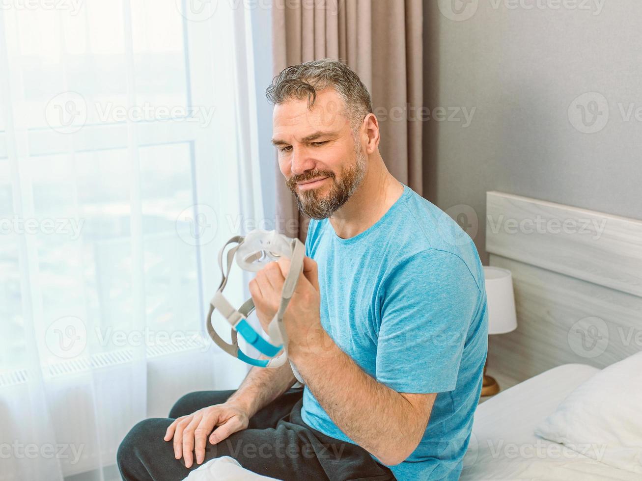 feliz homem descansado com problemas respiratórios crônicos depois de usar a máquina de cpap sentada na cama no quarto. cuidados de saúde, cpap, terapia para apneia obstrutiva do sono, conceito de ronco foto