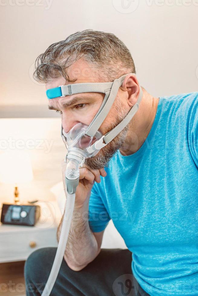 homem chocado infeliz com problemas respiratórios crônicos, surpreso com o uso da máquina de cpap sentada na cama no quarto. cuidados de saúde, cpap, terapia para apneia obstrutiva do sono, conceito de ronco foto