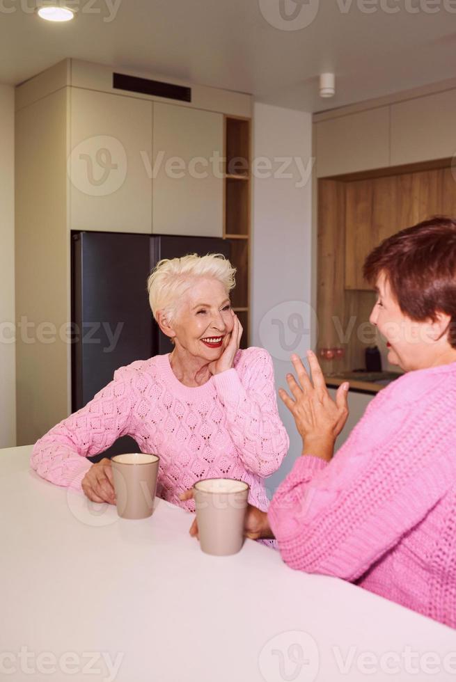 duas mulheres elegantes sênior em suéteres rosa sentadas com canecas na cozinha moderna fofocando. amizade, conversa, fofoca, relacionamentos, notícias, conceito de família foto