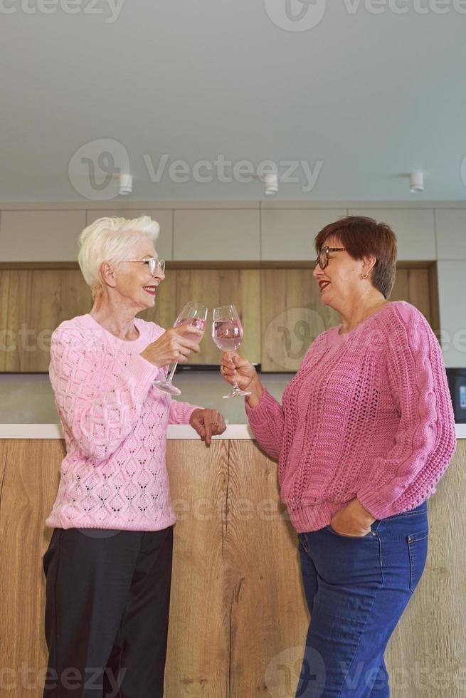 duas elegantes mulheres sêniors em suéteres cor de rosa bebendo vinho rosé na cozinha moderna fofocando. amizade, conversa, fofoca, evento, relacionamentos, notícias, conceito de família foto