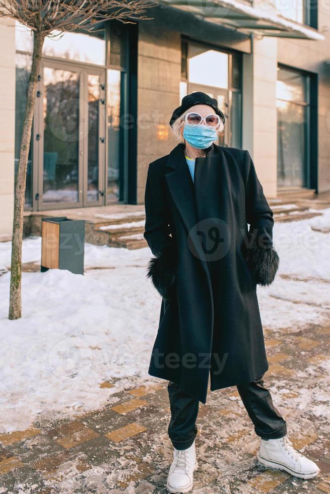 mulher elegante sênior na boina e elegante casaco preto e máscara médica, caminhar ao ar livre. bloqueio, pandemia, conceito de proteção foto