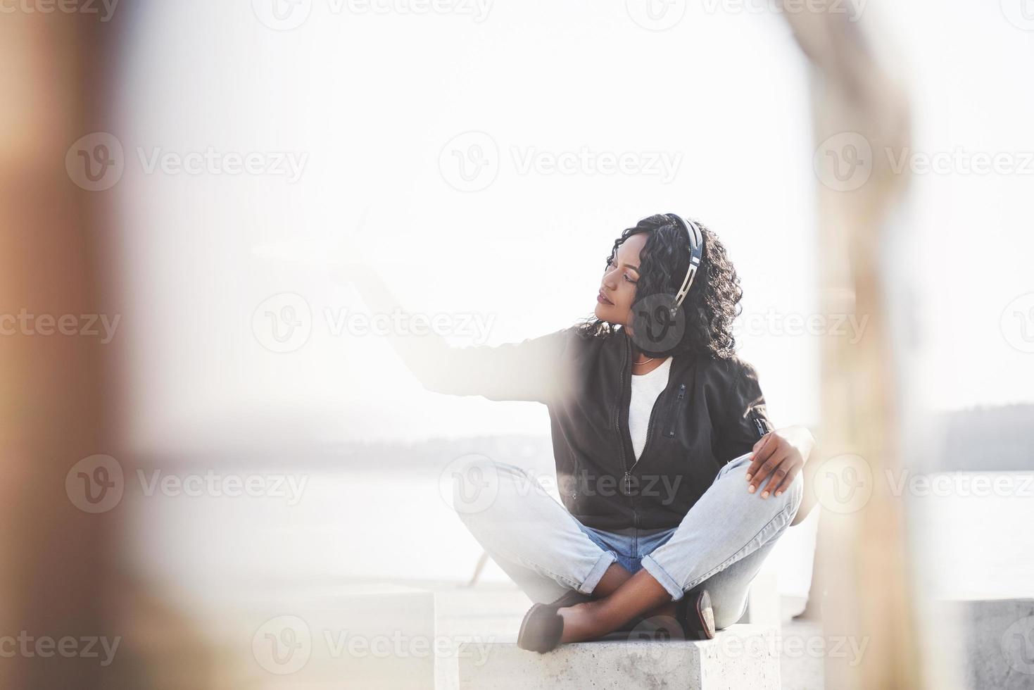 retrato de uma bela jovem afro-americana sentada na praia ou lago e ouvindo música em seus fones de ouvido foto