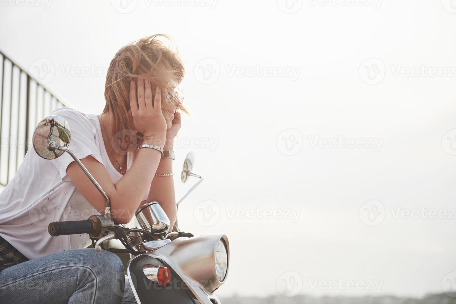 retrato de um hipster linda garota sentada em uma scooter retrô preta, sorrindo, posando e desfrutar do sol quente de primavera. foto