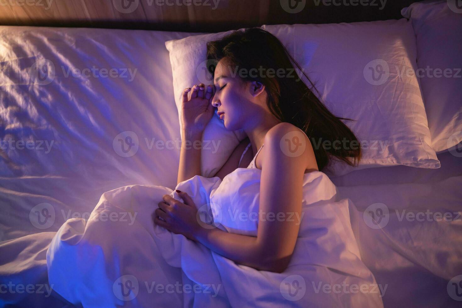 ásia mulher dormindo dentro confortável cama com sedoso roupa de cama às noite claro. mulheres deitado dentro cama e guardando olhos fechadas enquanto coberto com cobertor. conteúdo e pacífico, alegre e confortável. foto