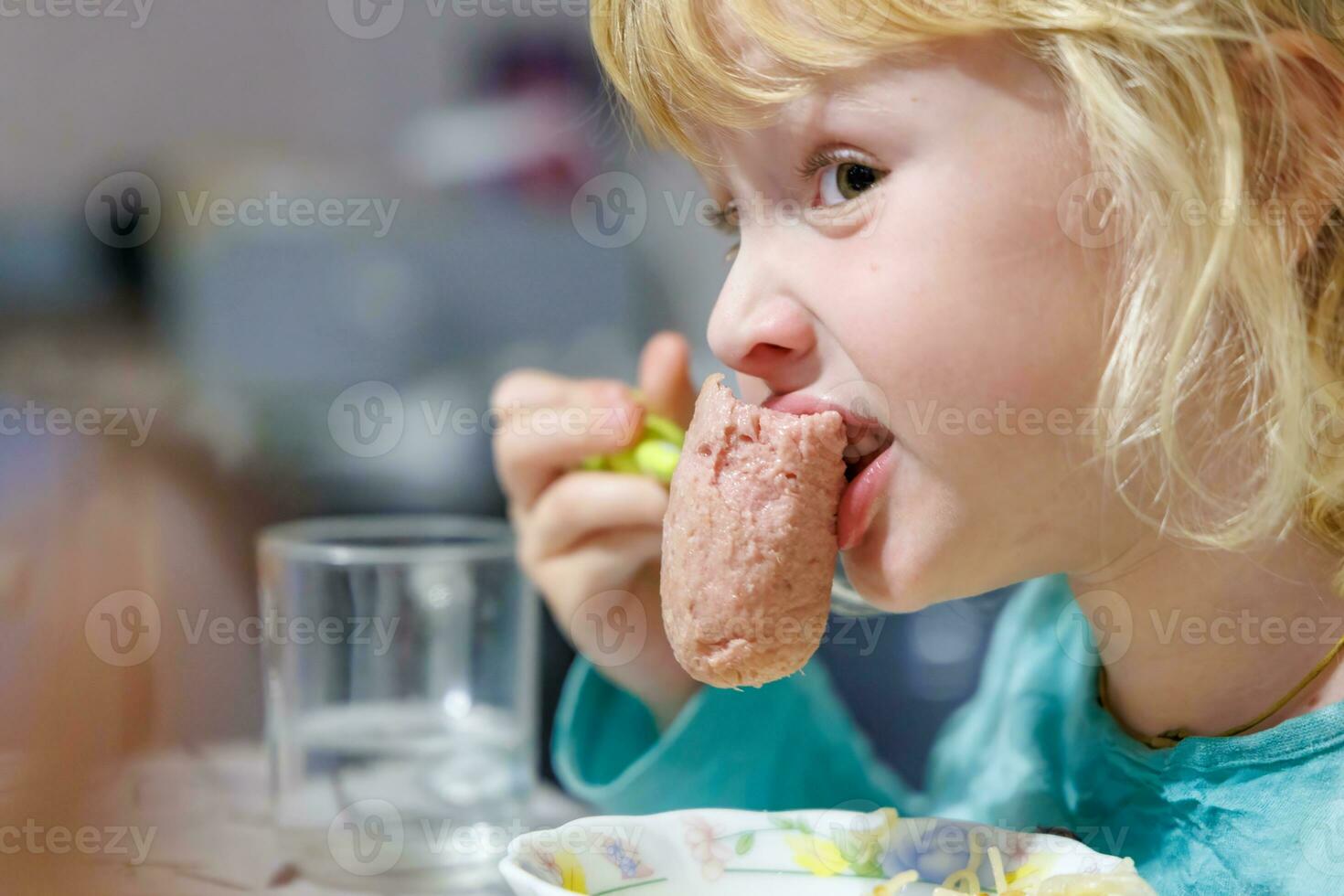 uma pequeno menina tem café da manhã às casa espaguete com salsichas. pequeno Loiras menina comendo jantar com garfo às mesa foto