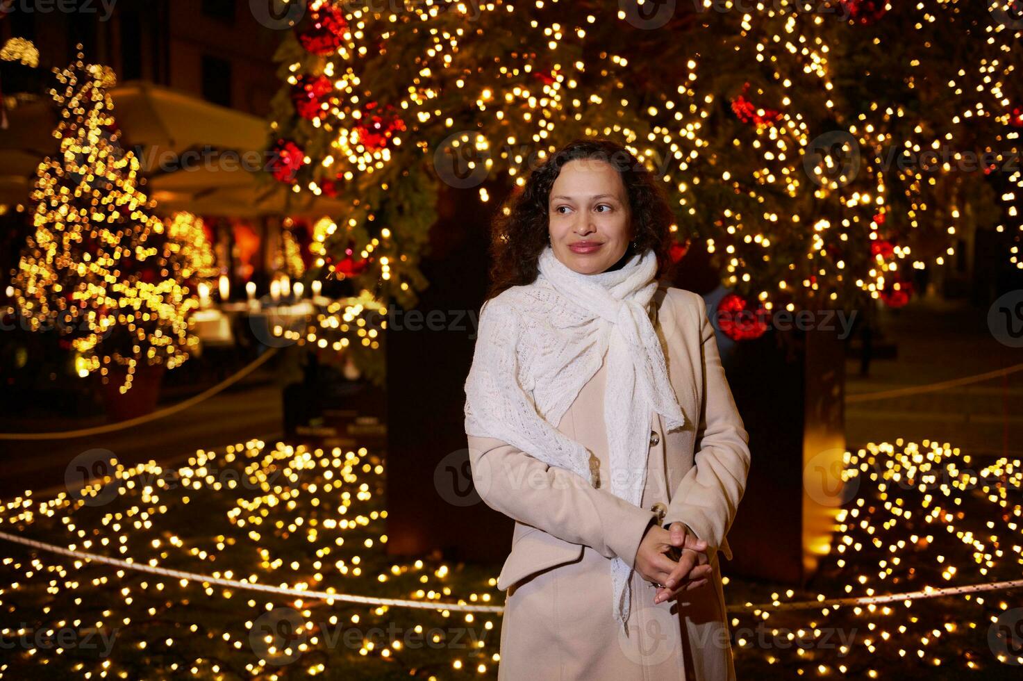 linda mulher goza a andar dentro Natal justo, admira lindo cidade rua decorado com festivo guirlandas às noite foto