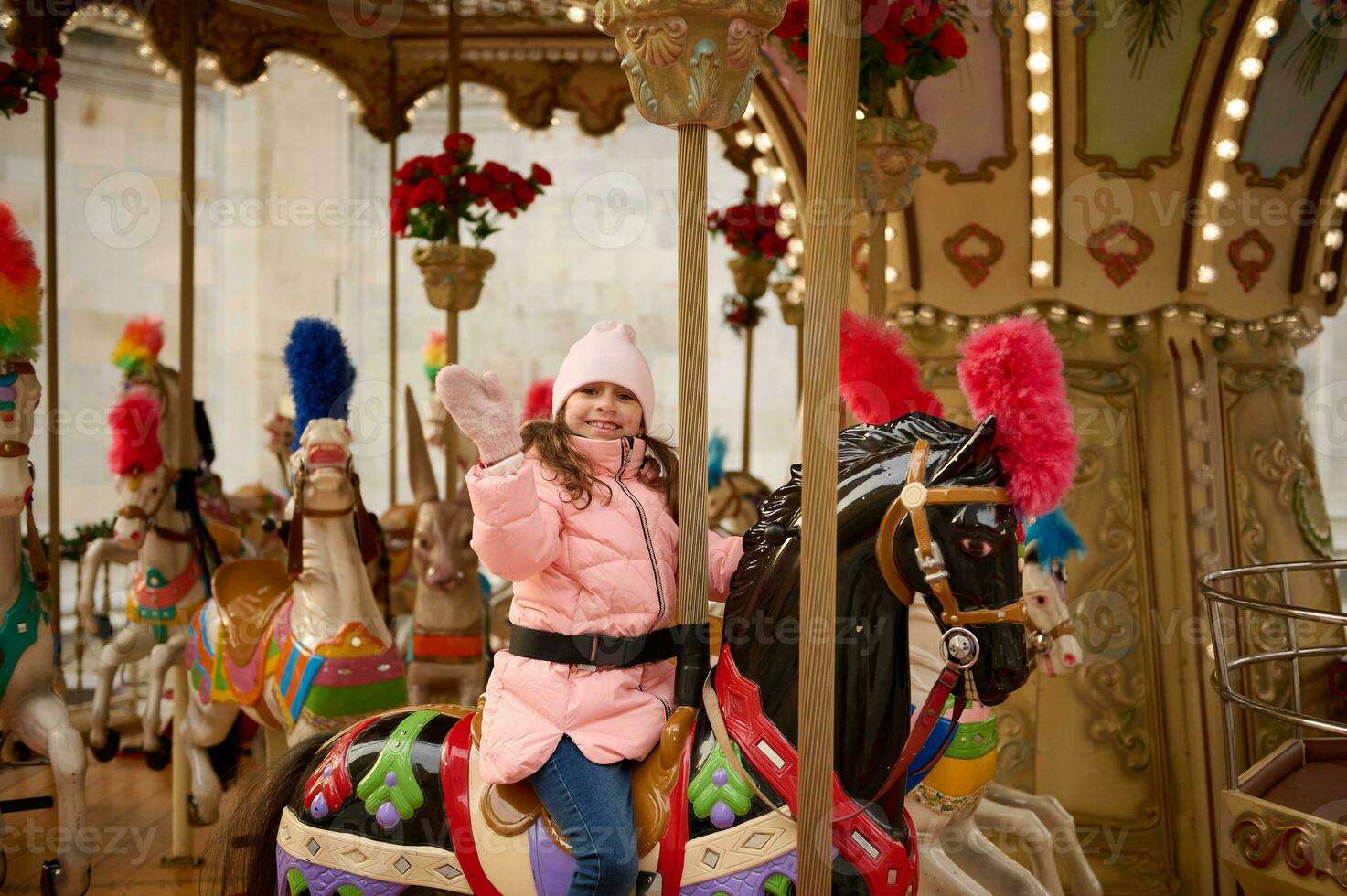 adorável sorridente criança menina acenando Olá enquanto equitação em alegre ir volta carrossel cavalo às Natal inverno mercado ao ar livre foto