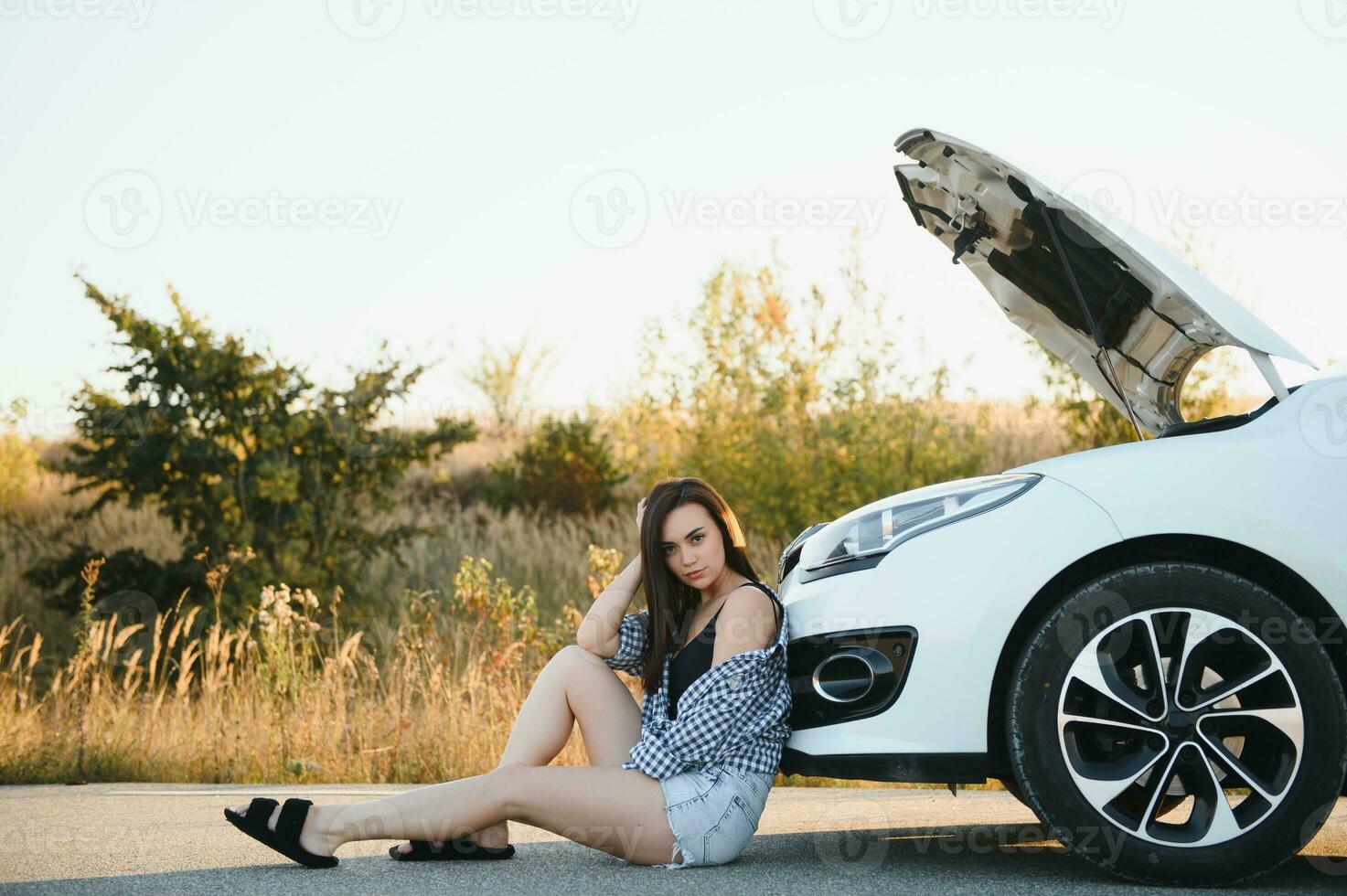 uma jovem menina senta perto uma quebrado carro em a estrada com a aberto capuz. foto