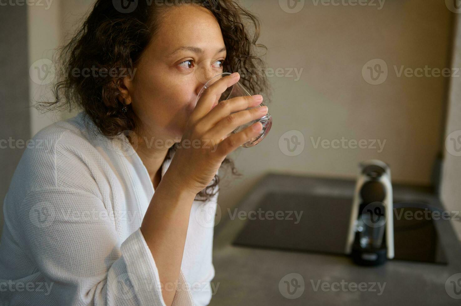 confiante multi étnico mulher vestido dentro branco manto, bebendo água enquanto levando medicamento, em pé de cozinha contador foto