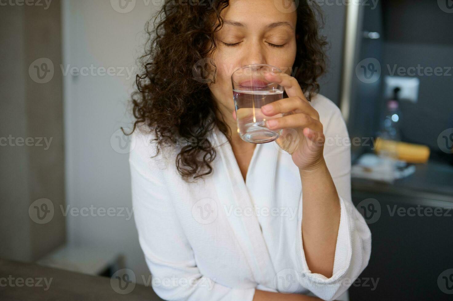 fechar-se autêntico retrato do uma multi étnico encaracolado morena mulher dentro branco manto, bebendo água enquanto levando remédio foto