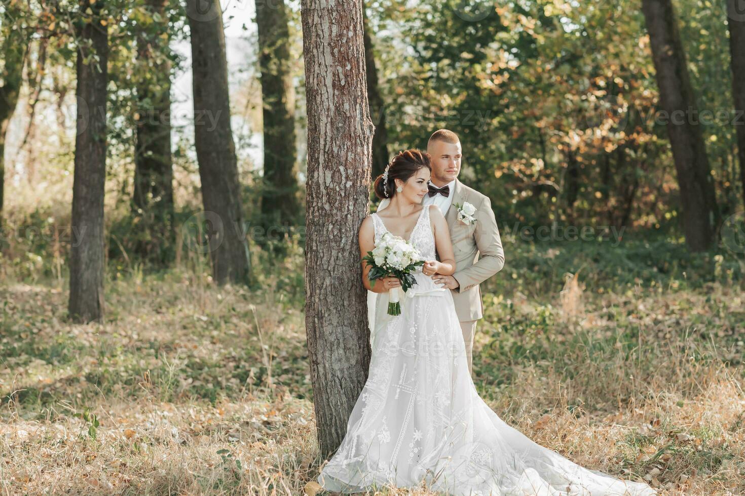 Casamento foto dentro natureza. a noiva e noivo estão em pé perto uma árvore sorridente e olhando ausente. a noivo abraços dele Amado a partir de atrás, a noiva detém uma ramalhete. retrato. verão Casamento