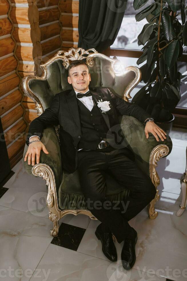 retrato do positivo jovem noivo dentro Preto terno com branco camisa sorridente e sentado em confortável Sombrio verde sofá dentro moderno hotel quarto em Casamento dia foto