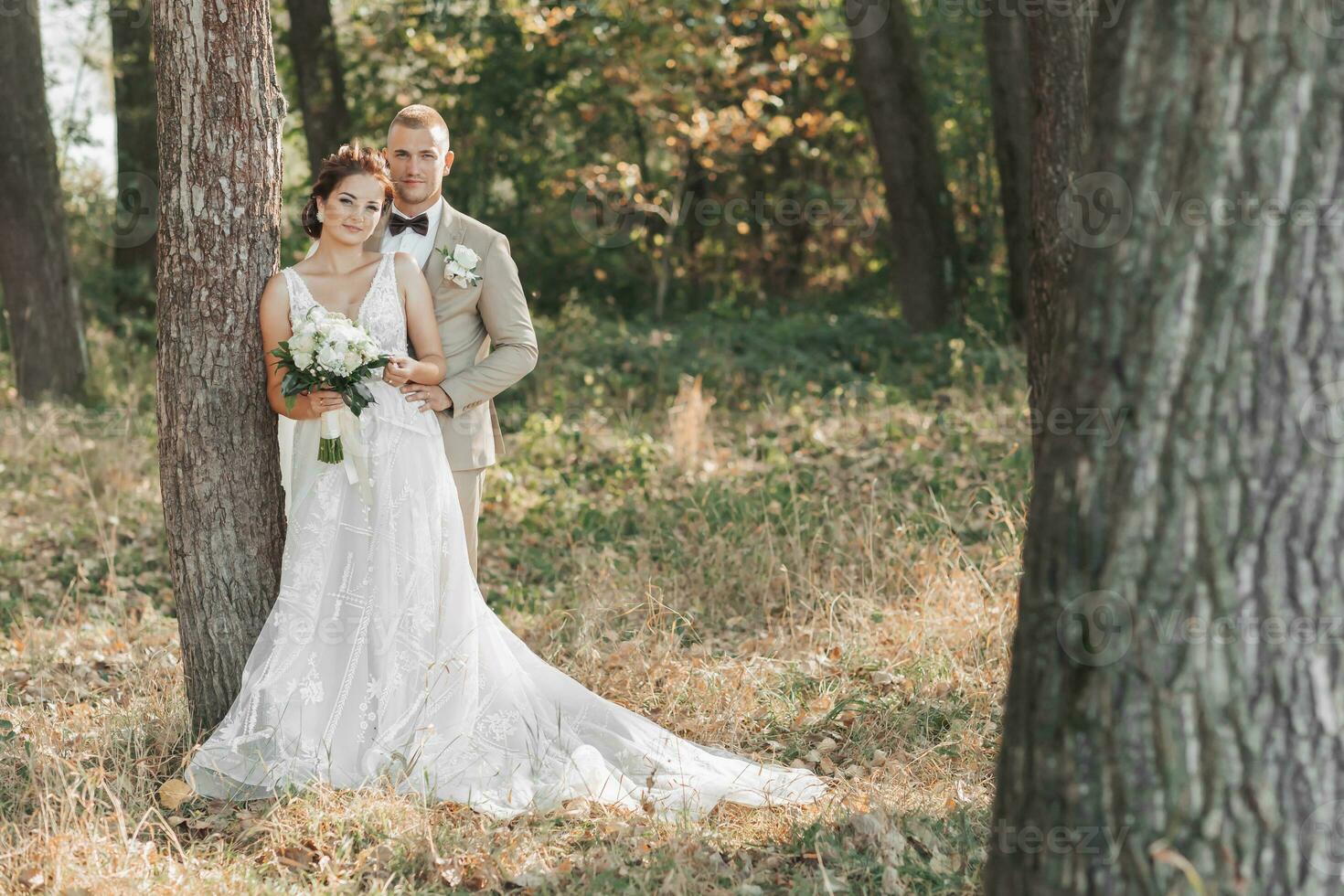 Casamento foto dentro natureza. a noiva e noivo estão em pé perto uma árvore sorridente e olhando às a Câmera. a noivo abraços dele Amado a partir de atrás, a noiva detém uma ramalhete. retrato. verão Casamento