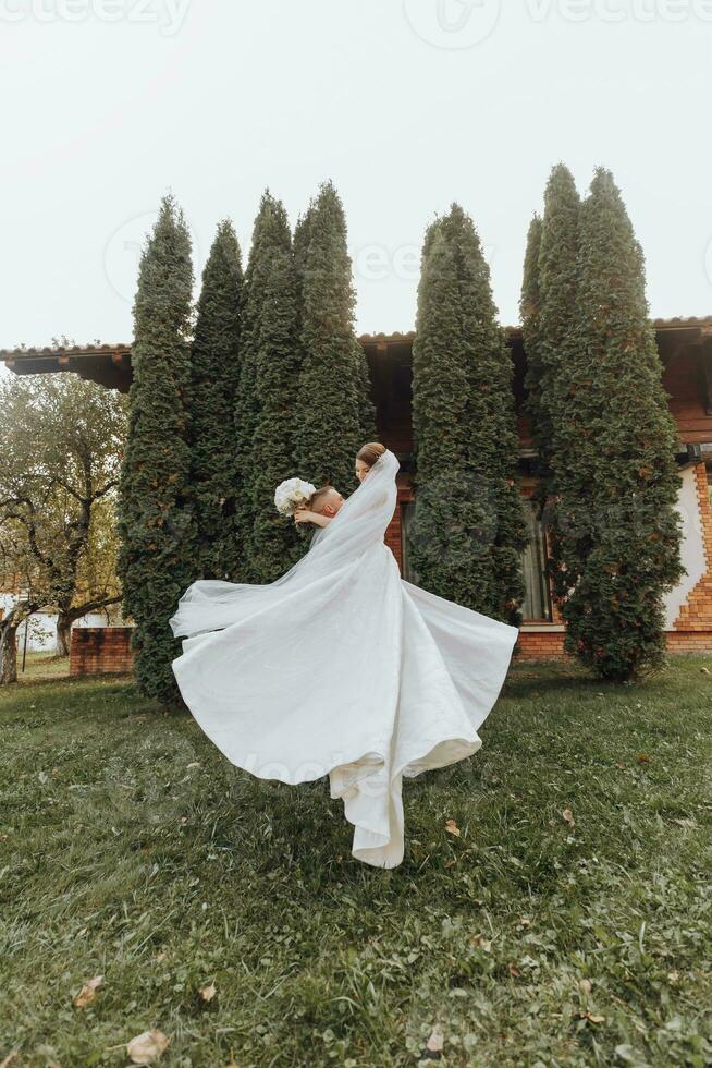 bonito noivo carregando uma lindo rindo noiva dentro dele braços dentro uma Primavera parque. a noivo é circulando com a noiva dentro dele braços foto