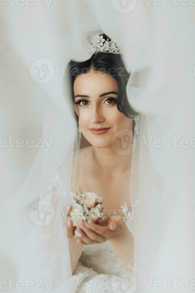 retrato. a morena noiva é em pé dentro uma chique coroa e branco vestir, embrulhado dentro uma véu, olhando baixa e cheirando a do noivo flor na lapela. linda Maquiagem e cabelo. foto