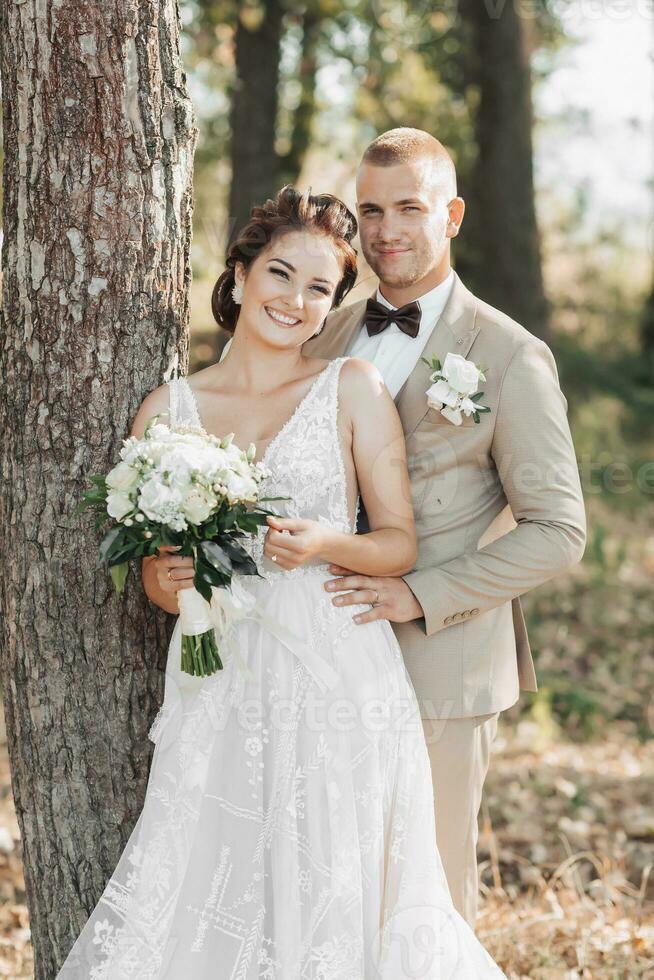 Casamento foto dentro natureza. a noiva e noivo estão em pé perto uma árvore sorridente e olhando às a Câmera. a noivo abraços dele Amado a partir de atrás, a noiva detém uma ramalhete. retrato. verão Casamento