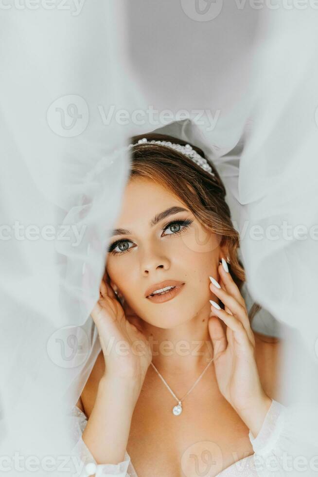 lindo encaracolado cabelos castanhos noiva dentro uma branco vestir poses para uma fotógrafo, em pé debaixo uma véu dentro uma lindo vestir com mangas. Casamento fotografia, fechar-se retrato, chique Penteado. foto