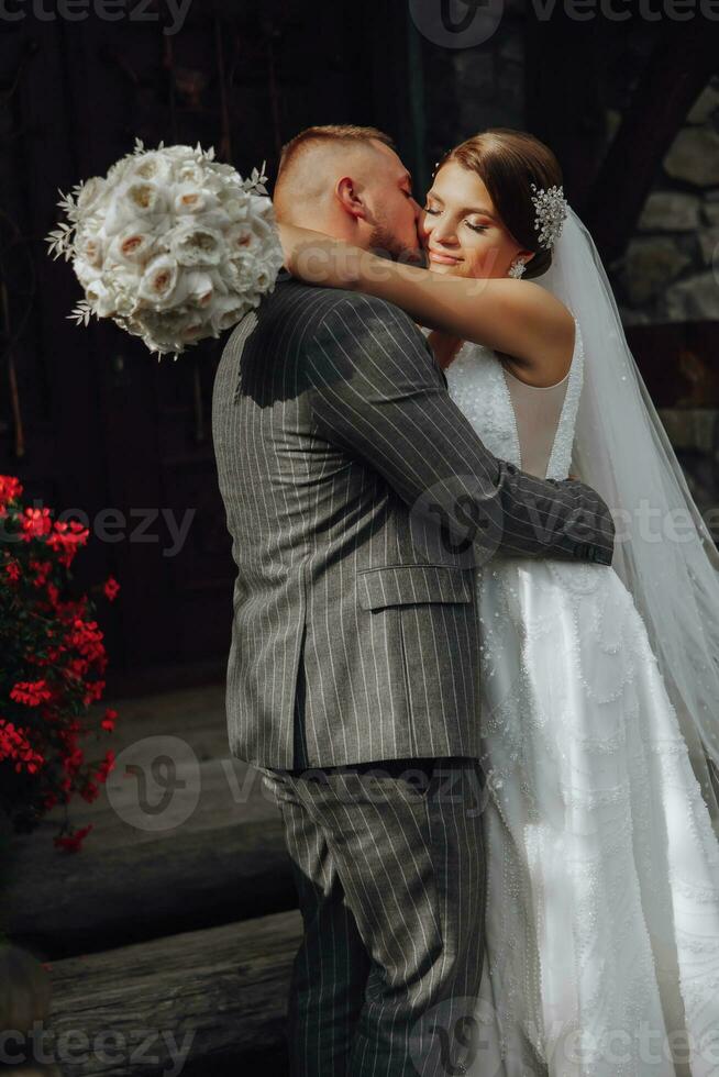 uma romântico noiva dentro uma branco vestir com uma ramalhete abraços a noivo perto a de madeira portão do a Igreja Entrada foto