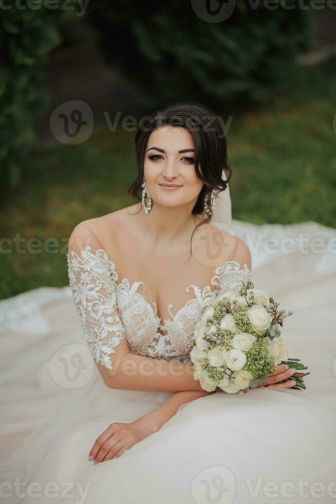 retrato do a noiva dentro natureza. uma morena noiva dentro uma branco renda vestir senta em a grama, poses, segurando uma ramalhete. lindo cabelo e inventar. aberto ombros. Casamento tiroteio foto