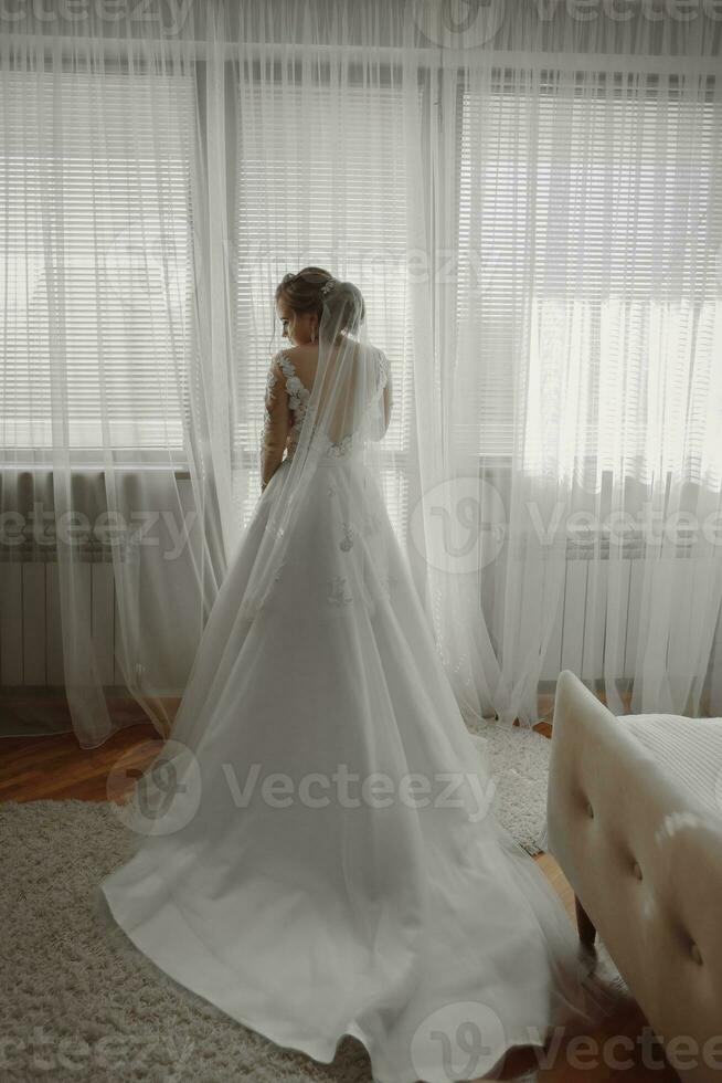 preparação para a casamento. lindo jovem noiva dentro branco Casamento vestir dentro de casa. luxo modelo parece às Casamento sapato, às casa dentro estúdio quarto com grande janela. a menina mostra uma foto a partir de a costas