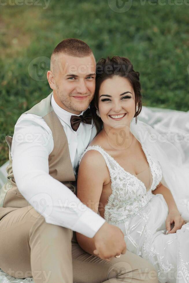 Casamento retrato do a noiva e noivo. feliz recém-casados estão sentado suavemente segurando mãos em a verde grama, posando. à moda noivo. lindo jovem noiva foto