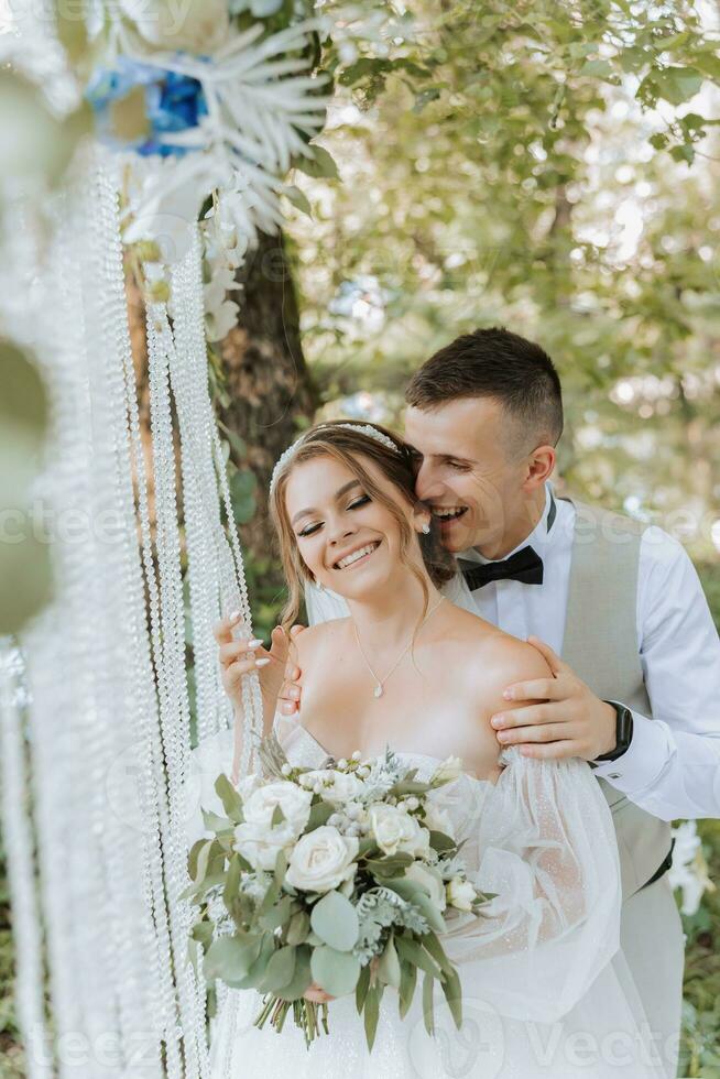 noivo e noiva em a Casamento dia, caminhando ao ar livre. a noivo abraços a noiva a partir de atrás de a ombros. pessoas estão feliz e sorriso. feliz casal dentro amor olhando às cada de outros foto