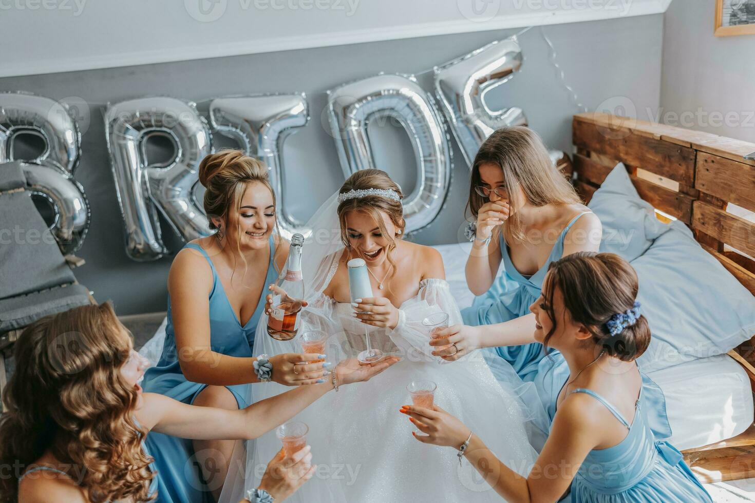 jovem damas de honra dentro azul seda vestidos beber champanhe dentro a da noiva sala. lindo mulheres a comemorar solteira festa sentado em cama e com champanhe. foto