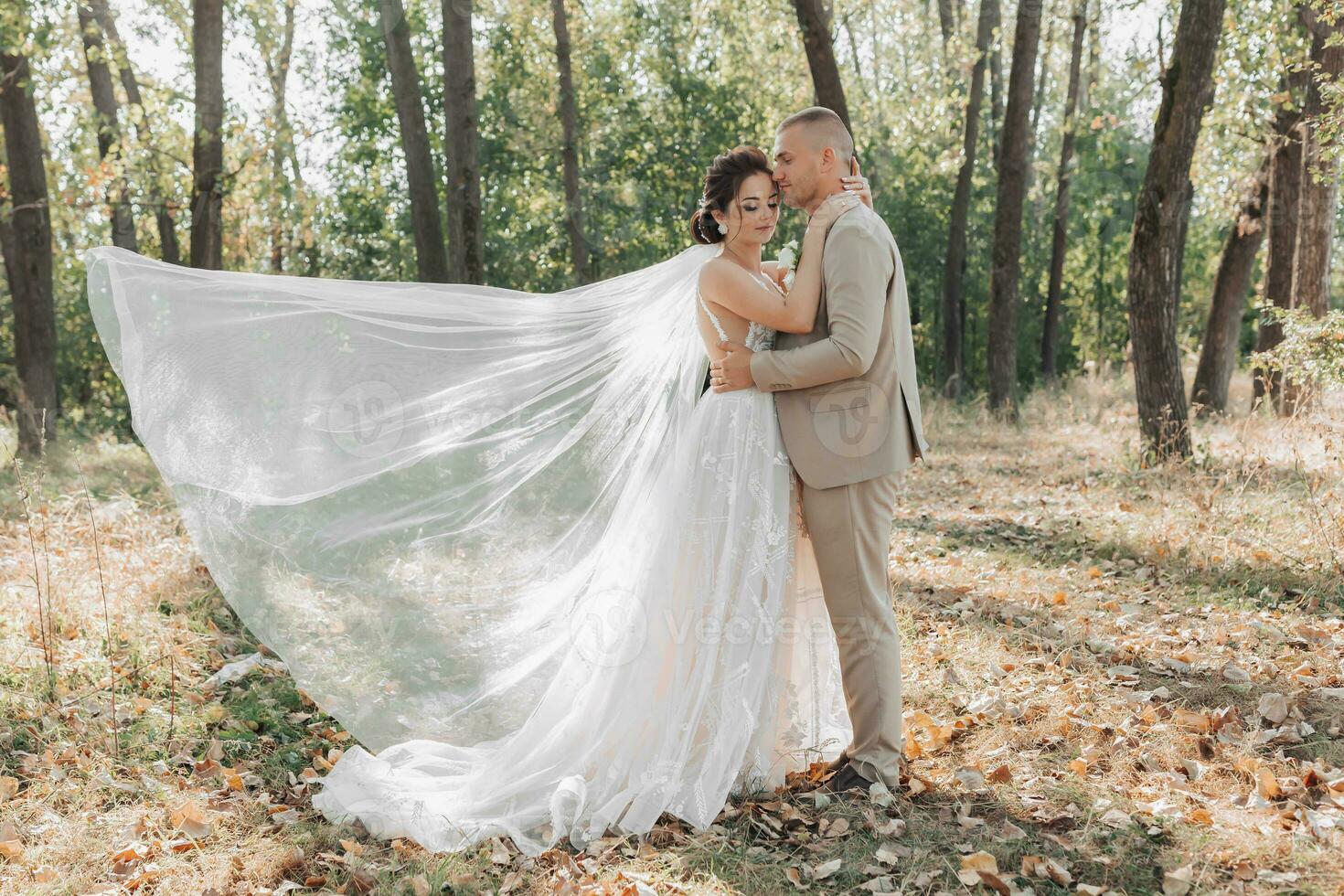 Casamento foto. a noiva e noivo estão em pé dentro a floresta abraçando, a noivo ternamente Beijos dele Amado em a testa. uma véu este cai belas dentro a ar. casal dentro amar. verão claro. foto