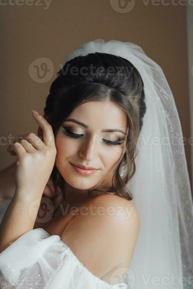 uma morena noiva dentro uma branco vestir detém dela linda cachos e parece abaixo. aberto ombros. lindo mãos. grandes véu. manhã do a noiva. foto