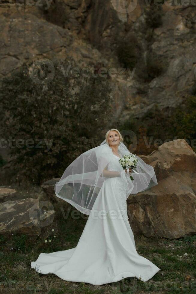 a noiva é em pé perto uma Rocha Alto dentro a montanhas, Próximo para uma ampla pedra. Casamento vestir. a vento lágrimas a véu foto
